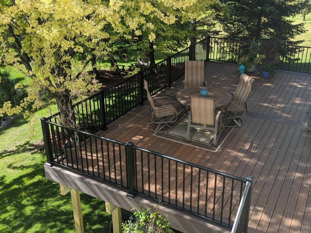 A large raised backyard deck with a table and chairs is surrounded by lush foliage.