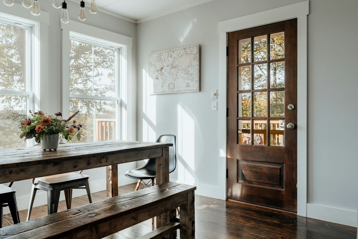 A brown, glass-paneled wooden door opens into a cozy dining area with a rustic table and bench, metal chairs and a vase of flowers.
