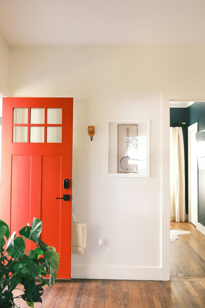 A red front door opens into a white foyer with hardwood floors and a monstera plant in the corner.