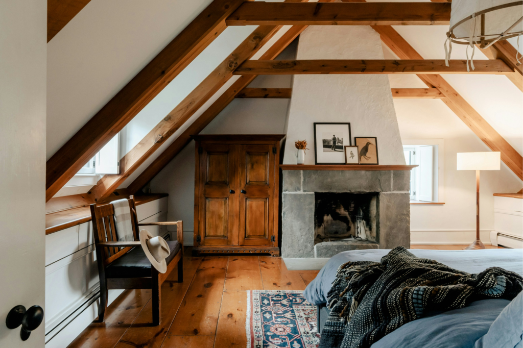 Cozy attic loft bedroom with exposed wooden ceiling beams, a bed and a warm hearth fireplace.
